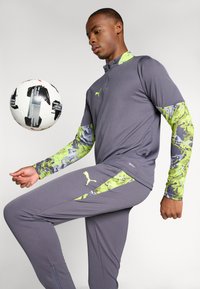 Gray sports shirt with long sleeves, accented in green and purple camouflage. Matching gray pants with green detailing. Soccer ball in focus.