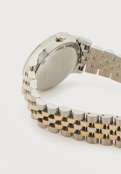 Μεταλλικό λουράκι ρολογιού από ασήμι και χρυσό με κούμπωμα και κορώνα, υπό γωνία που εμφανίζει την πίσω πλευρά της θήκης του ρολογιού και τους κρίκους σε λευκό φόντο.