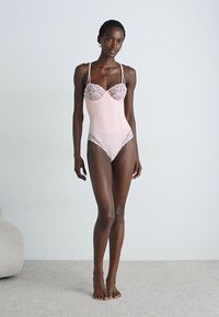 Femeie înaltă îmbrăcată cu un bodysuit roz pal cu dantelă, stând desculță pe un covor deschis, în fața unui perete alb simplu.