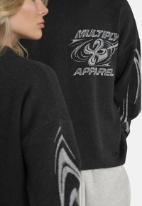 Suedou negru din tricot, cu un model de spirale albe și textul "MULTIPLY APPAREL" pe spate. Croi relaxat, cu umeri lăsați.