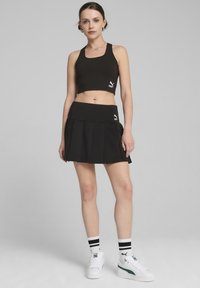Fekete cropped top és plisszírozott szoknya szett, az illeszkedő dizájn, puha anyag és fehér logó részletek jellemzik. A modellen fehér sportcipő és csíkos zoknik vannak.