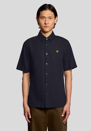 Mann trägt ein schwarzes kurzärmeliges Hemd mit Knöpfen und einem gelben Vogel-Logo auf der linken Brust, kombiniert mit braunen Hosen, steht vor einem grauen Hintergrund.