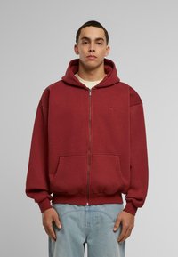 Maroon zip-up hoodie made of soft fabric, featuring a front pocket, ribbed cuffs, and a drawstring hood. Subtle branding on the chest.