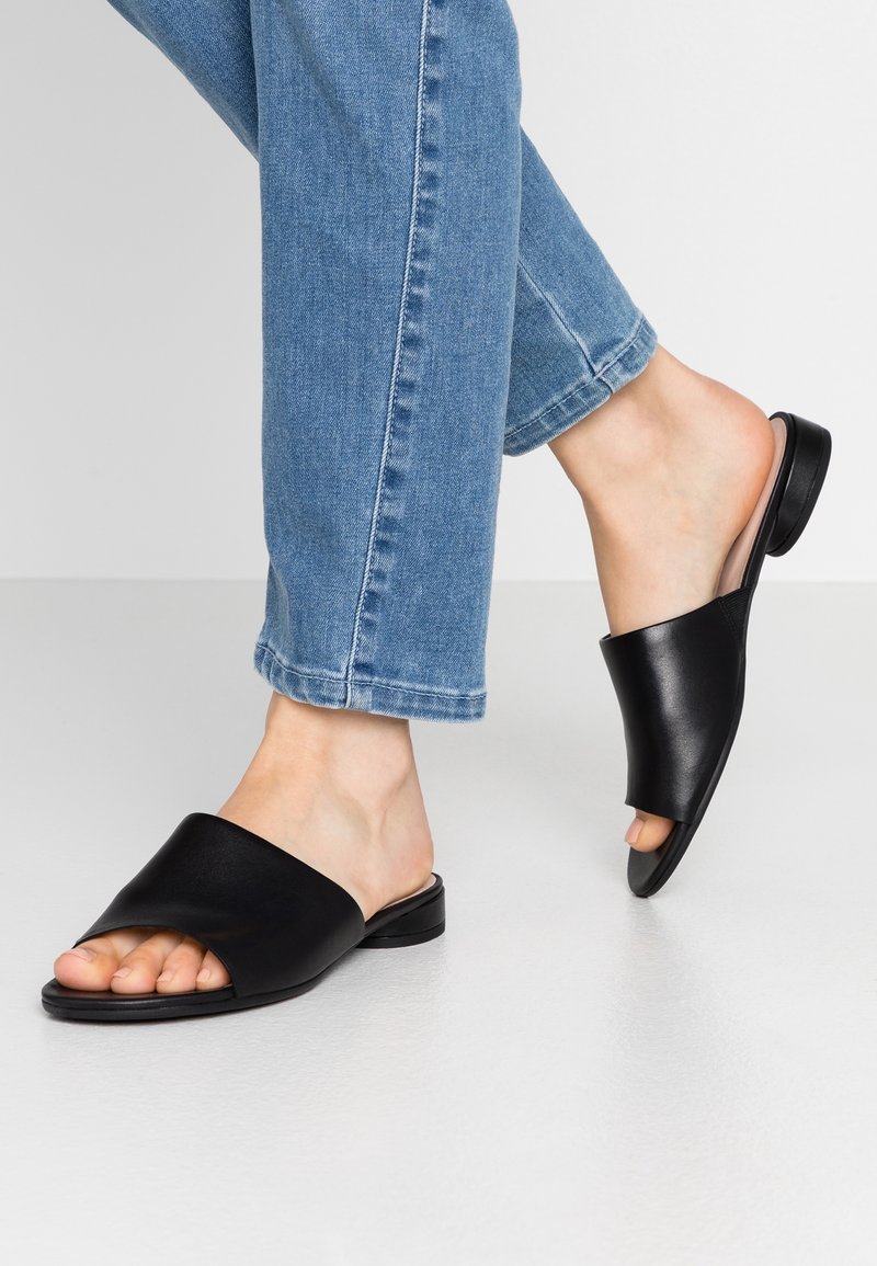 Černé kožené sandály s otevřenou špičkou a širokým páskem, plochou podrážkou, nošené s ohrnutými modrými džíny na neutrálním pozadí.