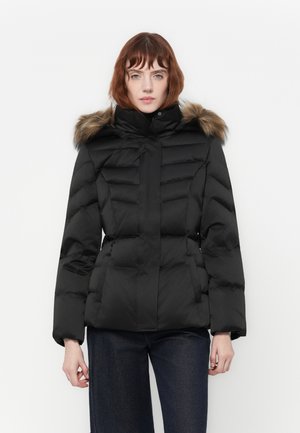 Femme portant une veste d'hiver noire matelassée avec une capuche à fourrure et un pantalon noir, se tenant devant un fond blanc uni.