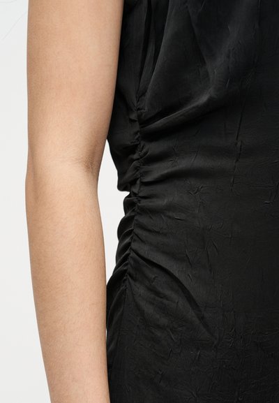 Κοντινή λήψη του βραχίονα ενός ατόμου δίπλα σε ένδυμα από μαύρο ρουχισμένο ύφασμα με ορατές ρυτίδες σε ουδέτερο φόντο.