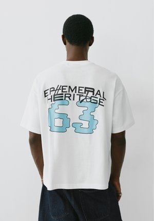 Personne aux cheveux courts portant un t-shirt blanc avec un texte noir et bleu "EPHEMERAL HERITAGE 63" dans le dos sur un fond uni.