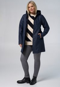 Marineblauwe waterdichte jas met een capuchon, drukknoopsluitingen en zijzakken, gedragen over een crèmekleurige trui met zwarte strepen. Grijze skinny jeans en zwarte laarzen.