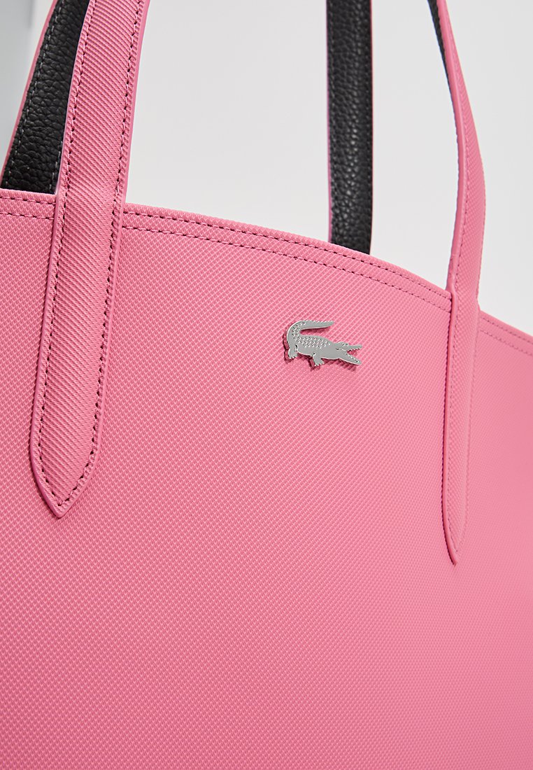 Rosa synthetische Einkaufstasche mit strukturierter Oberfläche, ausgestattet mit zwei Griffen und einem metallischen Lacoste-Logo-Akzent. Schwarzes Innenfutter sichtbar.