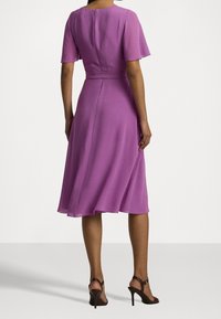 Fialové midi šaty s přiléhavým živůtkem, krátkými volánovými rukávy a rozšířenou sukní. Obsahují zipové zapínání a splývavou látku. Sandály na vysokém podpatku.