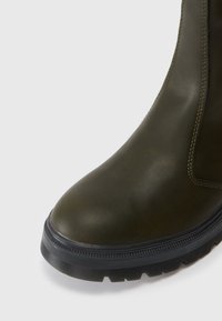 Bottine en cuir vert olive avec un bout arrondi, des panneaux latéraux élastiques et une semelle en caoutchouc noire avec un motif de semelle texturé.