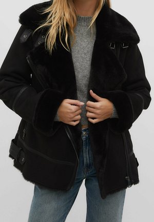 Personne portant une veste en peau retournée noire ouverte sur un pull en maille gris et un jean bleu, ajustant la veste avec les deux mains.