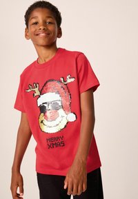 T-shirt en coton rouge avec un Santa Claus en sequins portant des lunettes de soleil, des cornes, et le texte "MERRY XMAS" en noir en dessous du motif.
