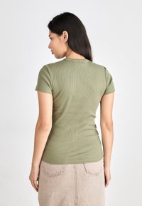 Olive green, ribbed short-sleeve t-shirt with a fitted design. The back features a round neckline and subtle texture, paired with a beige skirt.