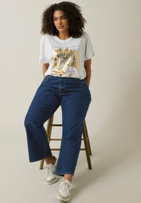 Camiseta gráfica blanca con diseño floral en dorado y negro, combinada con jeans de mezclilla azules de tiro medio. El modelo está de pie sobre un taburete de madera.
