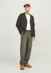 Cardigan marrone scuro con polsini e orlo a costine, t-shirt bianca, jeans larghi verde-blu, e stivali marroni. Cappellino rosso con dettaglio del logo.