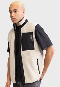 Gilet en polaire crème avec une poche noire, fermeture éclair à l'avant et tissu texturé. Porté par-dessus une chemise à manches courtes noire, avec des couleurs contrastantes.
