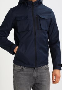 Marinblå jacka med huva, två bröstfickor och framdragkedja. Tillverkad av ett slätt, lätt material; har justerbara ärmslut.