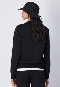 Sudadera negra de manga larga con puños acanalados, combinada con un sombrero tipo cubo negro. La tela parece suave y tiene un corte relajado. Camiseta blanca visible.