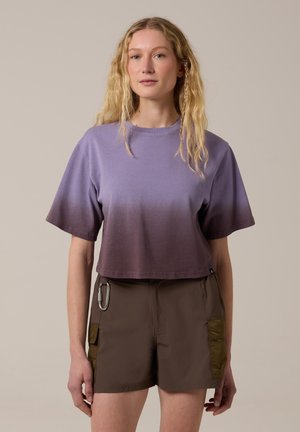Jonge vrouw met lang blond haar draagt een paars ombre cropped T-shirt en bruine korte broek met zijzakken en een karabijnhaak.
