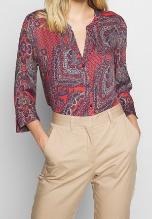 Blusa a righe rossa e blu con colletto a V, maniche a 3/4 e chiusura con bottoni, abbinata a pantaloni beige con tasche laterali.