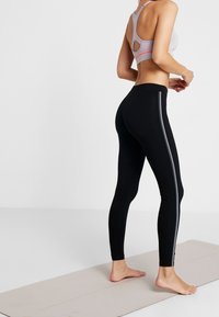 Höga svarta leggings med en silverrand på sidan, tillverkade av stretchmaterial. Matchas med en ljusgrå sport-bh med utskärningsdetaljer.