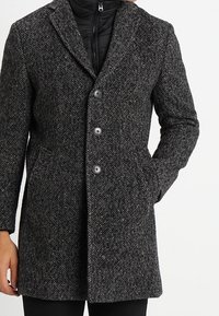 Man in een donkergrijze, gestructureerde wollen jas met drie knopen en een hand in zijn zak boven een zwarte, gewatteerde ritslaag.