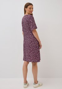 Kurzärmliges Kleid mit floralem Muster in Dunkelpurpur und Creme, mit A-Linie-Silhouette und weichem Stoff. Trägt weiße Sneaker.
