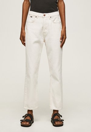 Personne portant un pantalon blanc taille haute à jambes droites et des sandales noires avec deux brides à boucle, debout devant un fond uni.