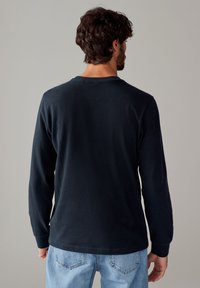 Camicia a maniche lunghe blu navy con scollatura a girocollo e polsini a costine, realizzata in morbido cotone, vista da dietro. Indossata con jeans azzurri chiari.