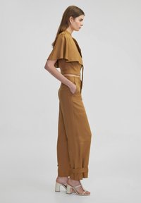Brown two-piece outfit with a cropped top featuring flutter sleeves and high-waisted trousers. The model wears strappy heels.
