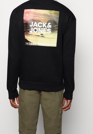 Osoba ubrana w czarną bluzę z nadrukiem zachodu słońca i białym napisem „JACK & JONES Established 1990” na plecach, połączoną z oliwkowymi spodniami cargo.