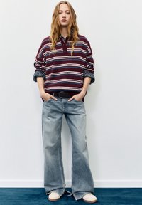 Pulover dungi vișinii cu guler cu nasturi, asortat cu blugi albăstri largi și adidași albi. Fundal neutru.