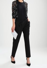Zwarte kanten bolero-jack over een zwarte top, gecombineerd met hoge zwarte broeken. Accessoires zijn een zilveren clutch en zwarte hakken.