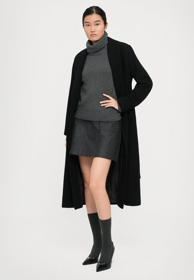 Woman wearing a black long coat, gray turtleneck sweater, dark gray skirt, matching socks, and black pointed heels standing with hand on hip.