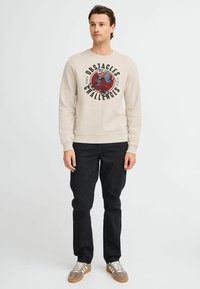Light beige sweatshirt with graphic text and a design centered on the front, paired with black pants and gray sneakers with white laces.