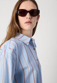 Light blue collared shirt with red and white vertical stripes, featuring a button closure. The person wears dark cat-eye sunglasses.