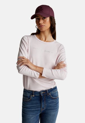 Vrouw in een roze langemouwen shirt met de tekst "United Colors of Benetton", blauwe jeans, een paarse pet met een doodskopembleem en ringoorbellen.