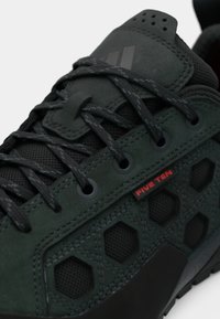 Κοντινό πλάνο μαύρου αθλητικού παπουτσιού Five Ten με εξαγωνικά πλέγματα στα πλαϊνά πάνελ και μαύρα κορδόνια με σχέδια που περνάνε μέσα από μεταλλικά τελείες.