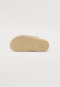 Beige skosula med ett texturerat vågmönster, försedd med en rektangulär logotyp på ena sidan. Utformad för förbättrat grepp och hållbarhet.