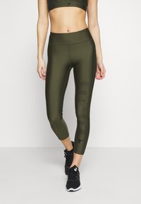 Olive green athletic leggings feature a high waistband, cropped length, and "WARRIOR" text down the side, made from a stretchy material.