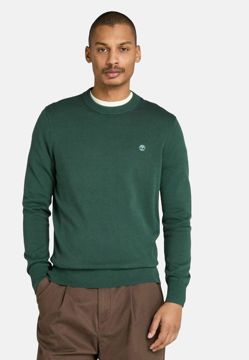 Ciemnozielony sweter dzianinowy z okrągłym dekoltem, ściągaczami przy mankietach i dolnym wykończeniu oraz małym logo na piersi, w połączeniu z brązowymi spodniami.