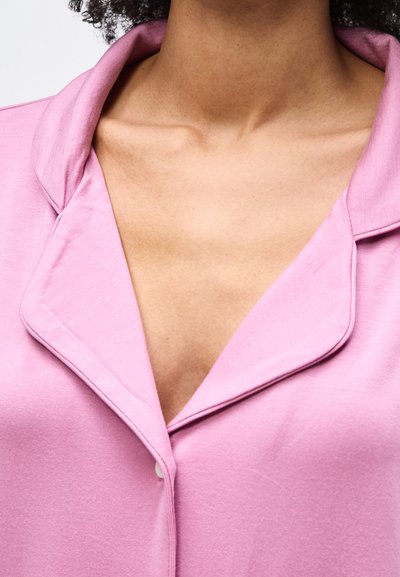 Κοντινό πλάνο ενός ατόμου που φοράει ροζ πουκάμισο με γιακά και ανοιχτό λαιμό, εμφανίζοντας το άνω μέρος του στήθους και τις κλείδες.