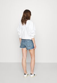 Camicia bianca oversized con bottoni, pantaloni shorts in denim blu strappati con orlo frangiato e sneakers bianche con dettagli neri, viste da dietro.
