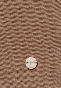 Barna kötött anyag sima textúrával, amelyen egy kerek, krémszínű gomb található, rajta az "Fynch Hatton" felirattal és egy fa logóval, 1998-ból.