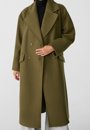 Femme portant un long manteau croisé vert olive sur un col roulé noir et un pantalon gris clair, sur un fond clair uni.