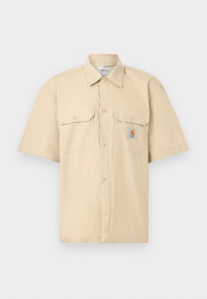 Μπεζ κοντομάνικο πουκάμισο με κουμπιά, δύο μπροστινές τσέπες με καπάκια, γιακά, και ένα μικρό σήμα με το λογότυπο Carhartt στην αριστερή τσέπη.