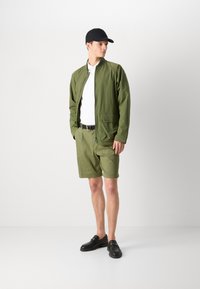 Zelená nylonová bomber bunda s předním zipem, doplněná o shodné šortky. Černá kšiltovka a mokasíny dokončují outfit, který se vyznačuje elegantním designem.