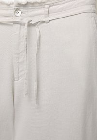 Gros plan sur un pantalon en lin blanc avec un bouton, des passants de ceinture et un cordon en tissu noué à la taille.