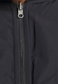 Svart zip-up-jacka med en slät yta, metallblixtlås med dragknapp och subtil broderad detalj på sidan.